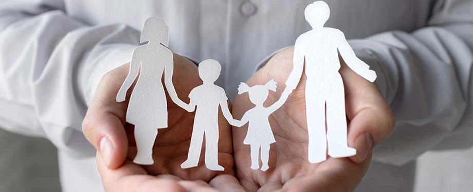 Qual o melhor seguro de vida familiar? Na imagem, mãos e figura de família representam coberturas oferecidas pelo seguro familiar, como morte e doenças graves